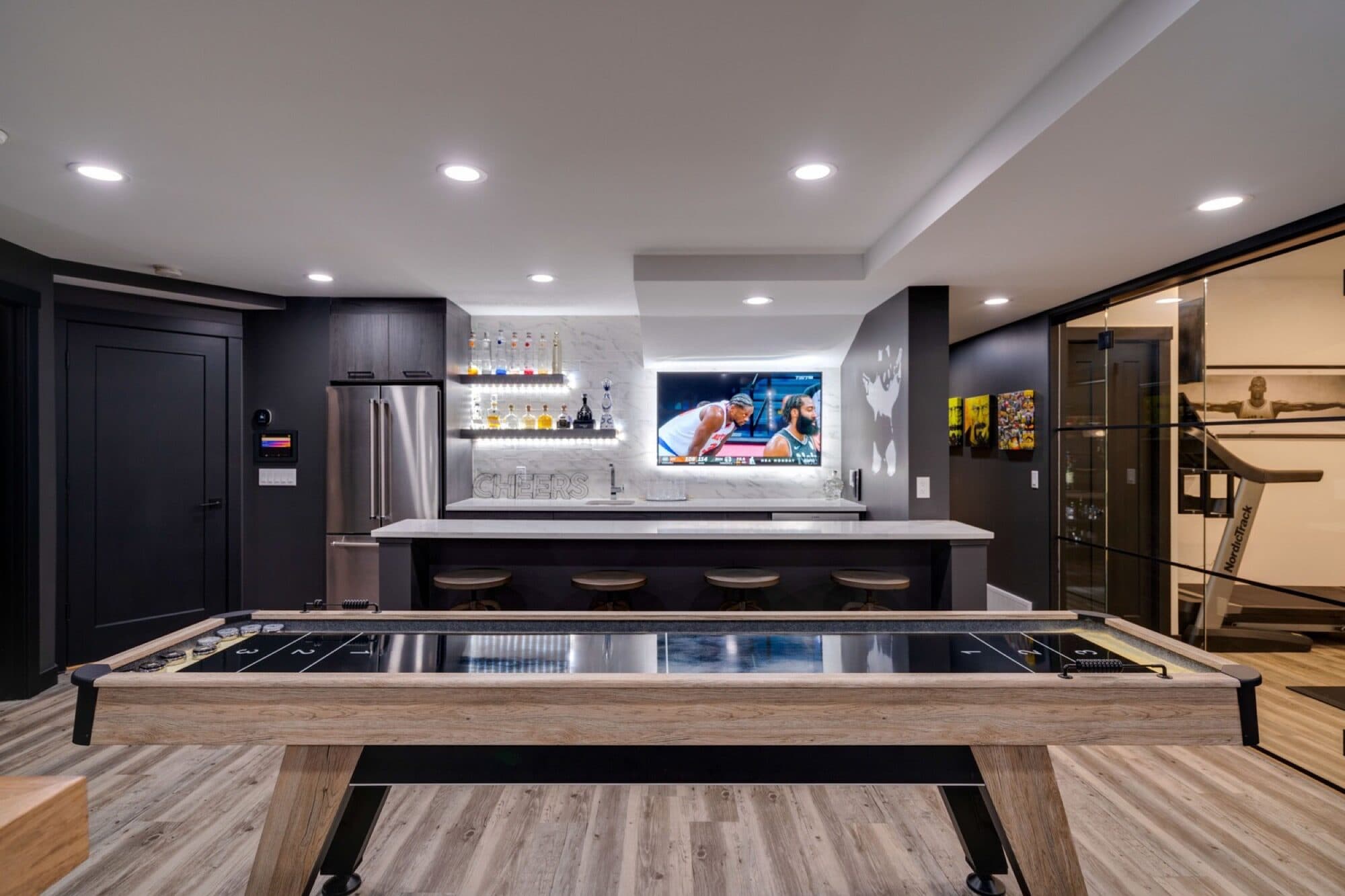 Basement rec room with shuffleboard, dark bar and gym glass wall