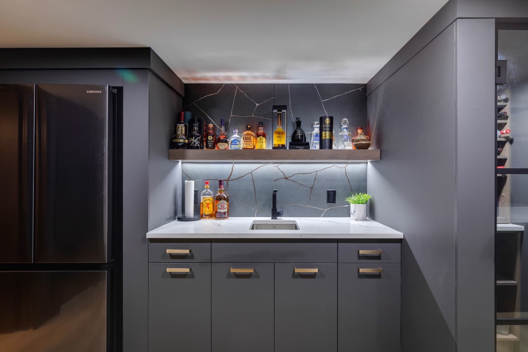 Basement wet bar with grey cabinets, marble backsplash and lit spirits shelf