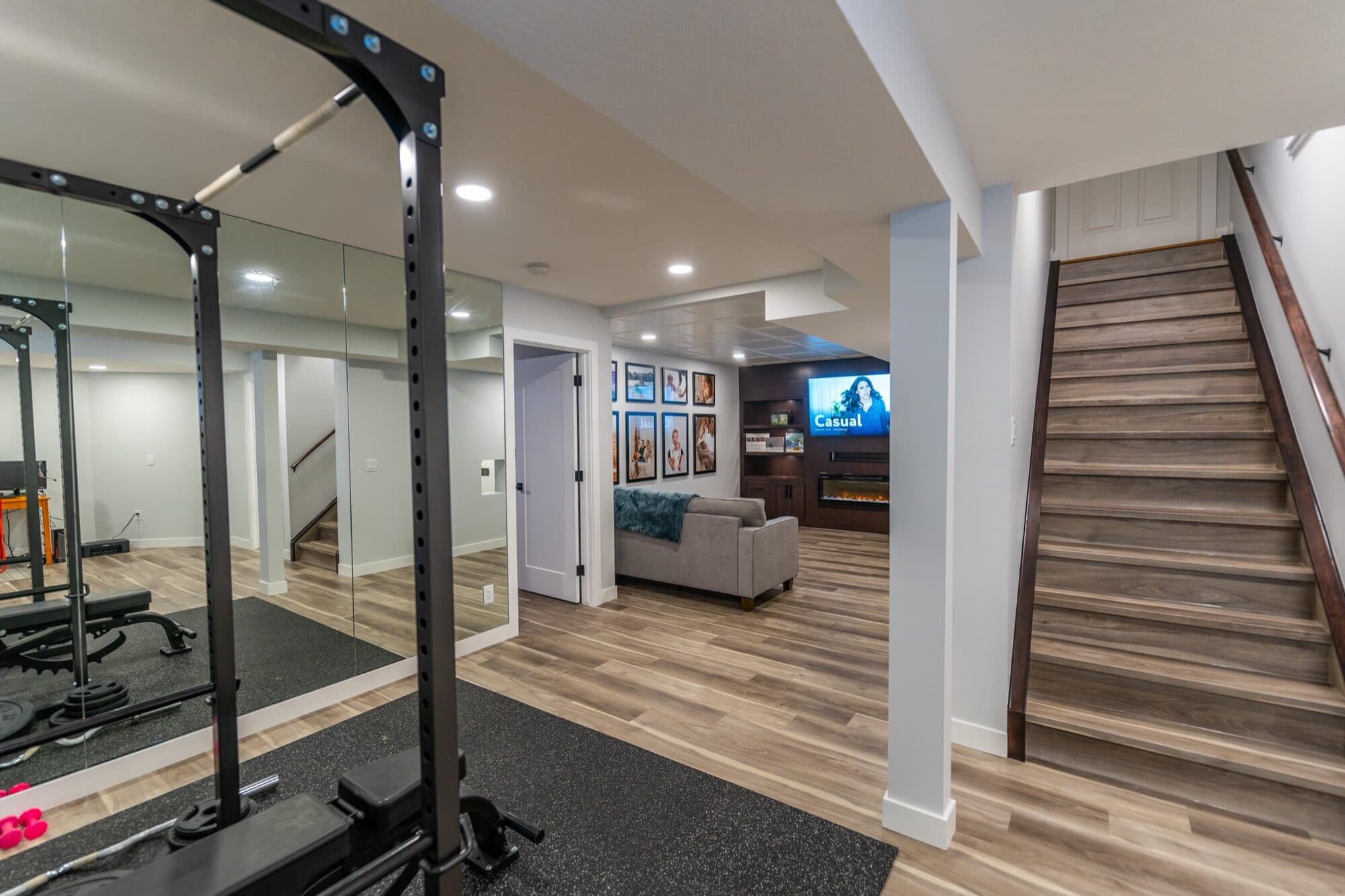Basement gym with power rack and mirror wall, rec room beyond