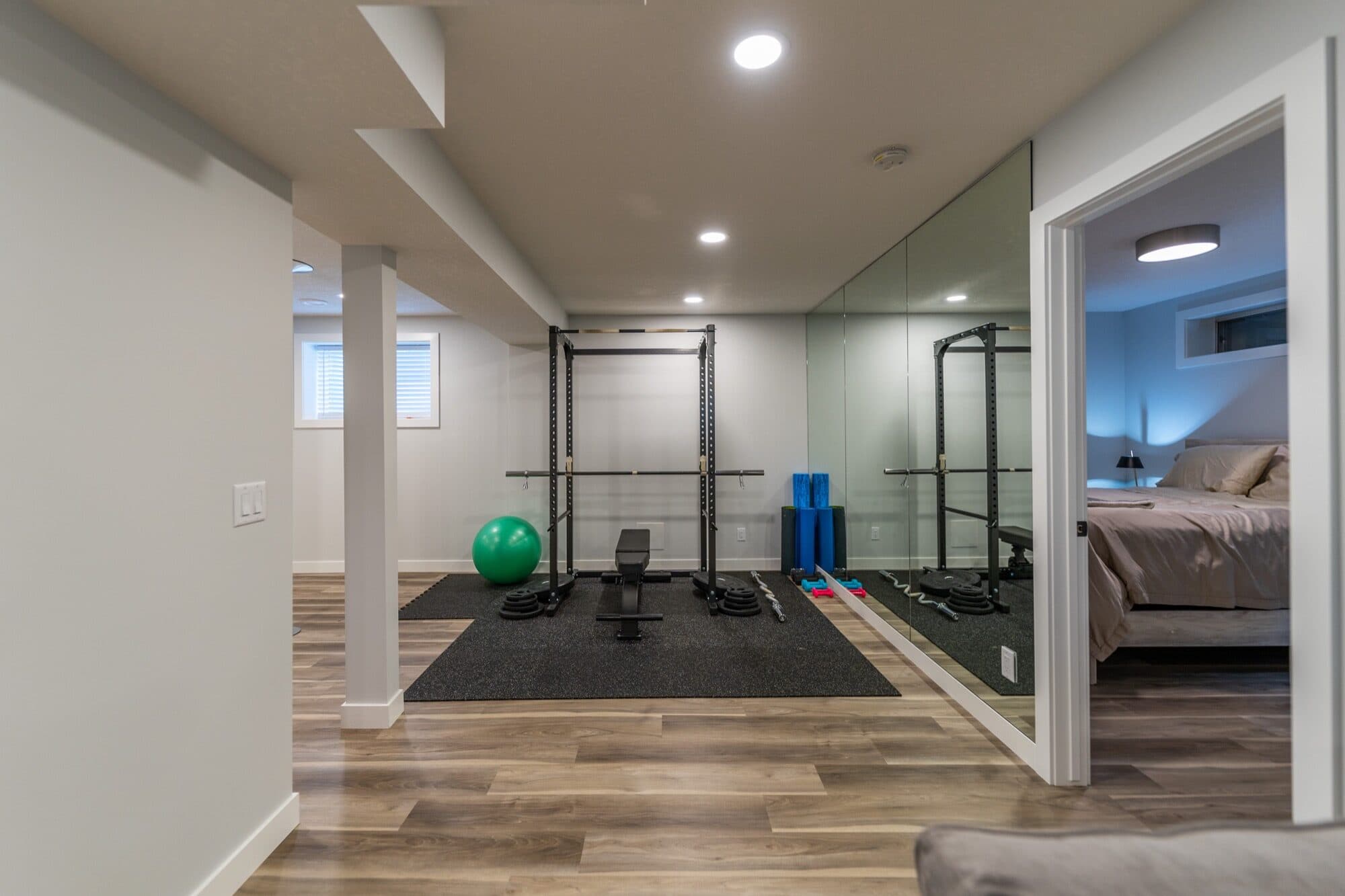 Basement home gym with squat rack, mirror wall and bedroom doorway