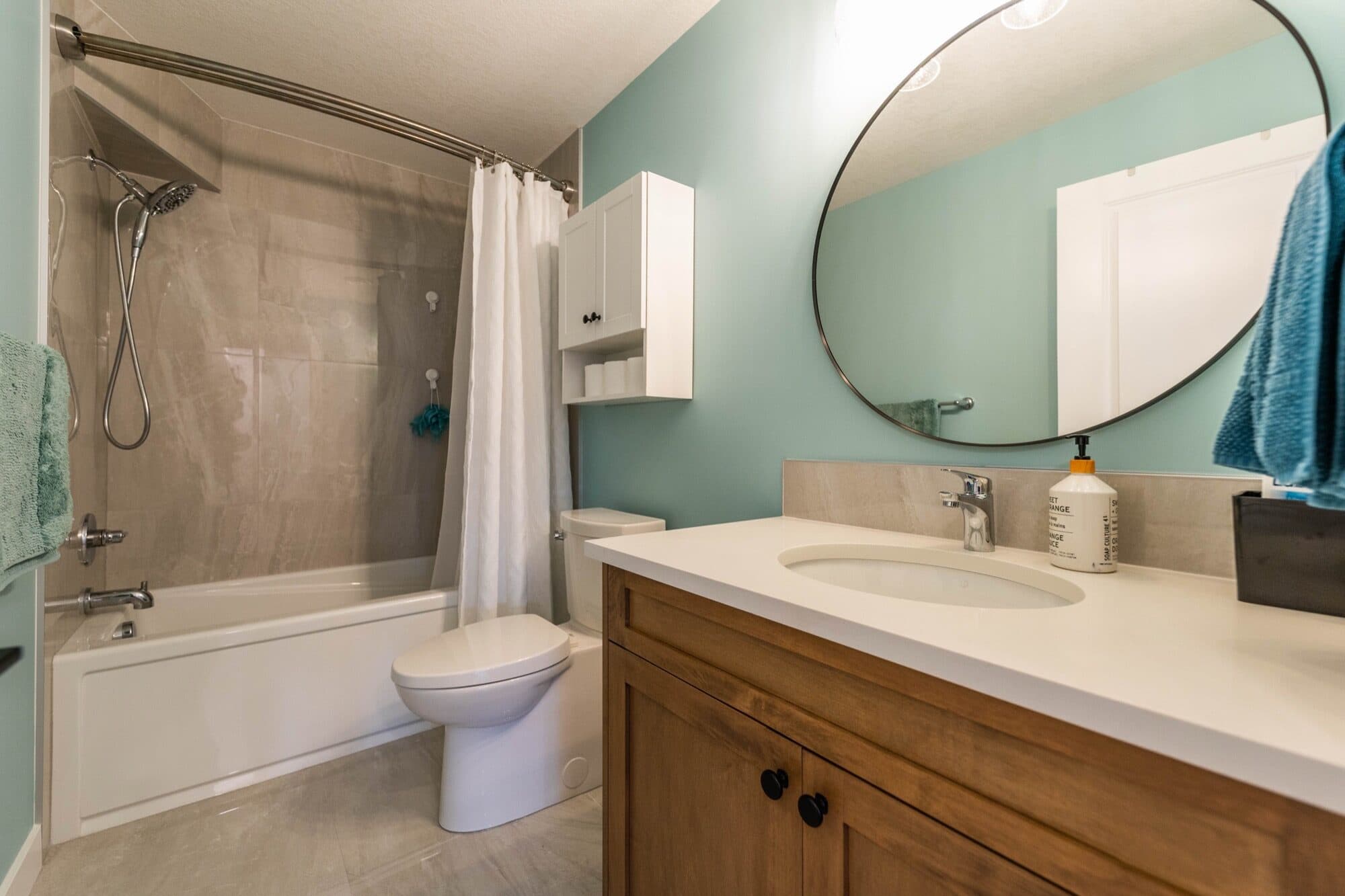 Basement bathroom with teal accent wall, round mirror, wood vanity and tub-shower